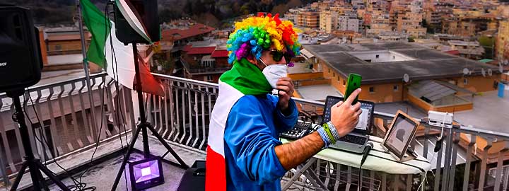 A DJ stands on a rooftop in Rome preparing to entertain his neighbors during the pandemic. The DJ wears a multicolored wig, sunglasses and a surgical mask. A laptop, microphone and other equipment sit on top of an ironing board in front of him. There are speakers and an Italian flag to his left. Buildings, trees and the sky are beyond the rooftop. 