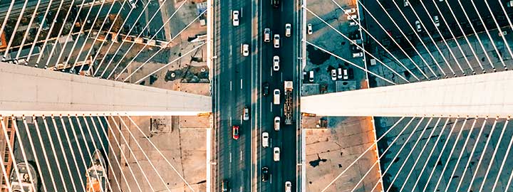 Aerial daylight view of Zolotoy Bridge in Vladivostok, Russia. In the center of the photo, vehicles appear tiny as they travel in six lanes of traffic across the bridge. The straight lines of the cables supporting the bridge rise from the lanes of traffic to the edges of the photo, forming geometric shapes. There are large ships in the water below the bridge. Cars on a road and in a parking lot, tree tops and buildings are also below the bridge.