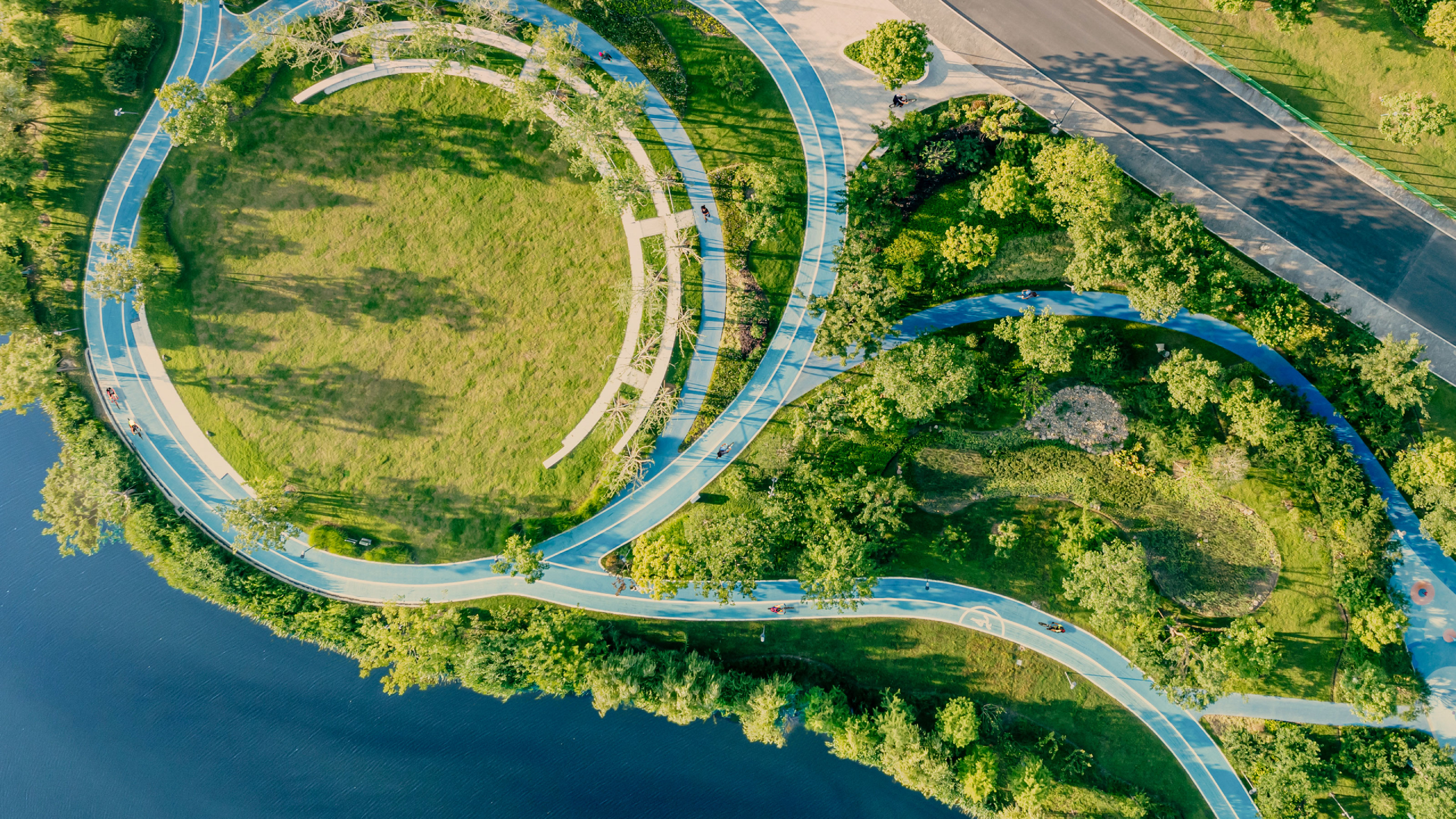 An aerial view of a city park at sunset in China. Paths wind gently through a verdant landscape, joining at the top left to form a circle enclosing an open, grassy space. Water is visible to the left, and a main road cuts through the upper right.